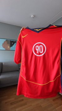Raudonas sportinis marškinėlis su V formos iškirpte, geltonais akcentais, balta apvali logotipu su „90“ ir Nike prekinio ženklo simboliu kairėje peties pusėje.