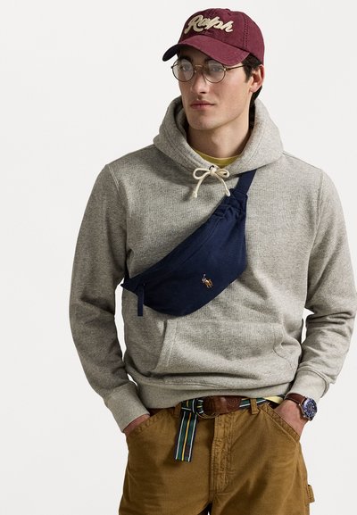 Navyfarbige Stoffschultertasche mit Reißverschluss, getragen über einem grauen Hoodie. Mit einer Kängurutasche, braunen Hosen und einem mehrfarbigen Gürtel. Runde Brille und Mütze.