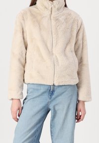 Faux fur jacket in beige with a high collar, front zipper, and long sleeves. Soft, textured material. Worn over light blue denim jeans.