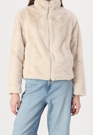 Faux fur jacket in beige with a high collar, front zipper, and long sleeves. Soft, textured material. Worn over light blue denim jeans.