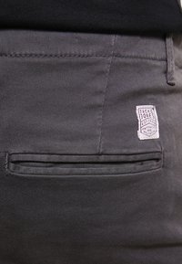 Ciemnoszare spodnie z gładkiej tkaniny, o tradycyjnym kroju, z boczną kieszenią i przyszytą metką z detalami marki.