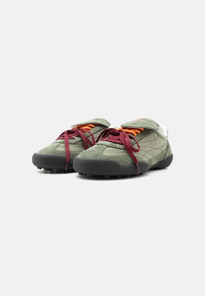 Paire de baskets en daim vert olive avec semelles noires et lacets rouges et orange sur fond blanc.