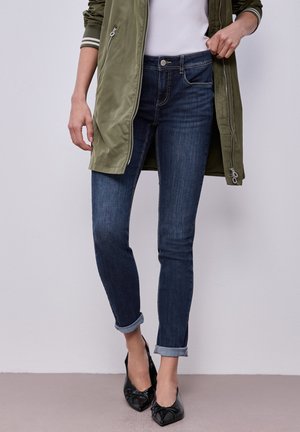 Osoba nosząca ciemnoniebieskie obcięte rurki, czarne spiczaste baleriny, biały top i zieloną długą kurtkę z detalami na zamkach.