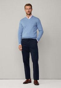 Hellblauer V-Ausschnitt-Pullover über einem rosa Kragenhemd, kombiniert mit dunkelblauen Hosen und braunen Lederschuhen. Sanfte Texturen und maßgeschneiderte Passform.