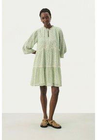 Rochie verde cu imprimeu floral, cu mâneci lungi și decolteu în formă de cheie, având volane în straturi și o croială lejeră, asortată cu sandale bej.