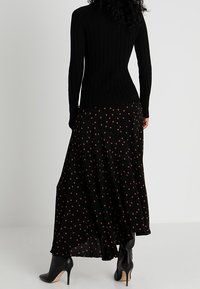 Schwarzes, geripptes Langarmoberteil kombiniert mit einem fließenden schwarzen Rock mit braunen Punkten. Schwarze, hochhackige Stiefeletten runden das Outfit ab.