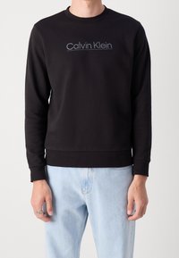Felpa nera in cotone con scollo rotondo, polsini e orlo a costine. Presenta il logo ricamato "Calvin Klein" in un colore a contrasto.