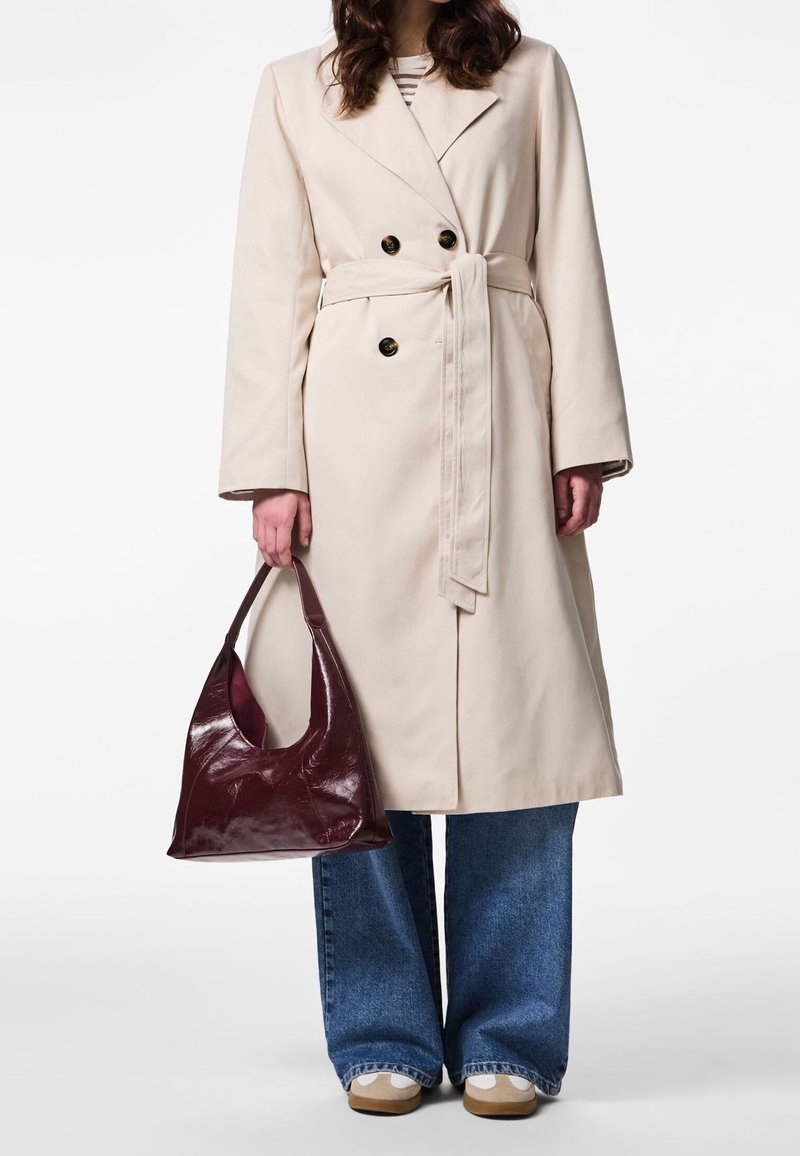 Femme portant un trench-coat beige ceinturé, un jean bleu à jambes larges, des baskets beiges, et tenant un sac à main en cuir rouge foncé dans sa main gauche.