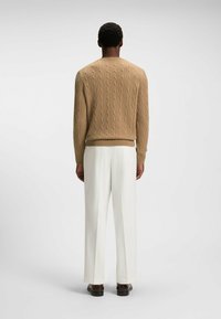 Beige Kabelstrickpullover mit Rundhalsausschnitt, kombiniert mit weißen weiten Hosen und braunen Schuhen, von hinten dargestellt.