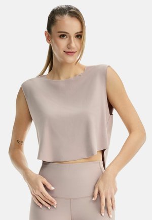 Donna con coda di cavallo che indossa un crop top beige senza maniche, largo, e leggings coordinati a vita alta, in posa con le mani vicino ai fianchi.