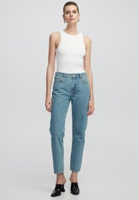Top blanco de canalé, jeans de tiro alto azul claro y zapatos negros tipo slip-on. La modelo está de pie con las manos a los lados contra un fondo sencillo.