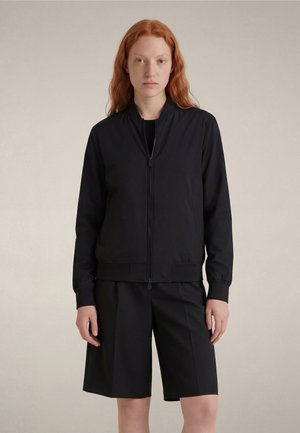 Młoda kobieta o długich rudych włosach, ubrana w czarną kurtkę na zamek i czarne spodenki do kolan, stoi na jednolitym tle.