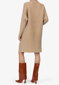 Robe en tricot côtelé beige à coupe décontractée, avec des manches longues et un col haut. Associée à des bottes mises hautes en croûte marron.