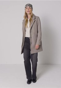 Ljusgrå kappa med skräddarsydd silhuett, knäppningar och två sidfickor, bärs över en vit topp och svarta jeans. Leopardmönstrad mössa.