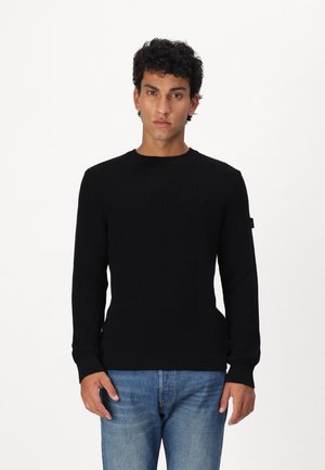 Schwarzer gerippter Strickpullover mit Rundhalsausschnitt und langen Ärmeln, ergänzt durch einen taillierten Saum. Kombiniert mit hellblauen Jeans.