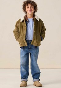 Veste en velours côtelé vert olive avec un col en fausse fourrure, superposée à une chemise bleu clair, assortie à un jean bleu clair et des chaussures beiges.