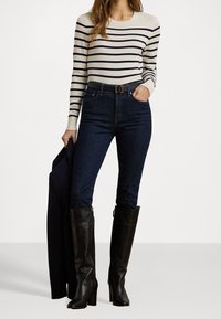 Pruhované krémové a tmavě modré tričko s dlouhým rukávem, spárované s vysokým pasem tmavých džínů a vysokými černými koženými botami na podpatku, držící černou bundu.