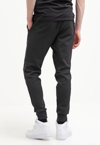 Man som bär svarta joggerbyxor med sidofickor och vita Nike-sneakers, står mot en vit bakgrund och tittar bort.