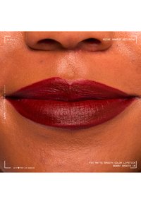 Gros plan sur des lèvres avec un rouge à lèvres mat couleur baie foncée sur un teint moyen-foncé, montrant une application lisse et uniforme ainsi qu'une texture naturelle de la peau au-dessus des lèvres.
