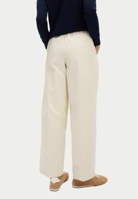 Pantalons larges crème avec une texture lisse et une taille élastique, associés à des baskets marron clair. La vue de dos montre un ample tombé.