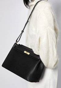 Sort læderhåndtaske med en glat tekstur, gyldent hardware-detalje og justerbar strop. Har en lynlåslomme foran og en rektangulær form.