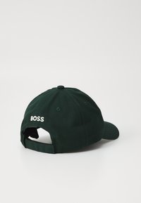 Mørk grønn baseballcaps med buet skygge, laget av bomull. Har hvitt brodert "BOSS"-logo bak og en justerbar stropp.
