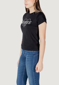 Zwarte fitted t-shirt met een hartenontwerp voorzien van rhinestones en het woord "GUESS" in vette letters, gecombineerd met blauwe denim jeans.