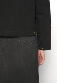 Schwarze Canvas-Jacke mit goldenen Beschlägen, sichtbaren Nähten und seitlichem Knopfdetail, kombiniert mit dunklen Jeans, die eine Gesäßtasche haben.