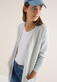 Cardigan grigio chiaro realizzato in materiale morbido, con apertura frontale, maniche lunghe e tasche laterali, abbinato a una t-shirt bianca e un cappellino blu.