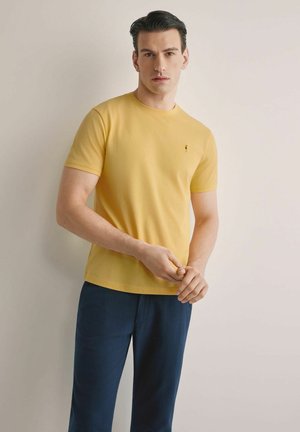 Mężczyzna ubrany w żółtą koszulkę z krótkim rękawem i ciemnoniebieskie spodnie, stojący na tle jednolitego jasnego tła z rękami złożonymi.