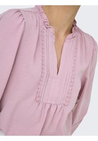 Blouse rose clair en tissu texturé, dotée d'un col à volants, d'une patte de boutonnage ornée de dentelle et de manches froncées pour une coupe ample.