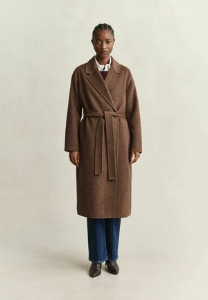 Femme portant un long manteau marron ceinturé, un jean bleu et des chaussures foncées, debout devant un fond clair uni.