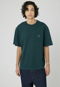 T-shirt en coton oversized vert forêt avec un col rond et un petit motif de champignon sur la poitrine gauche. Manches courtes et coupe décontractée.