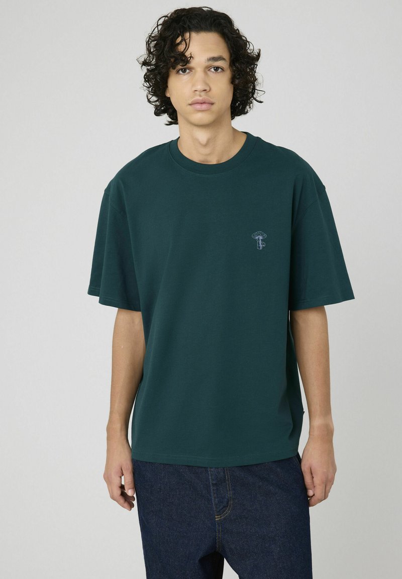 T-shirt en coton oversized vert forêt avec un col rond et un petit motif de champignon sur la poitrine gauche. Manches courtes et coupe décontractée.