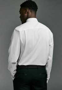 Camicia bianca a maniche lunghe con colletto classico e polsini con bottoni. Indossata con pantaloni neri. Tessuto liscio e vestibilità sartoriale.