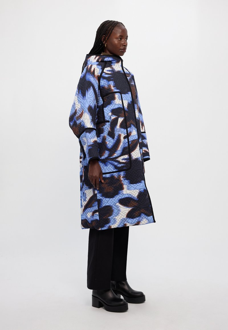 Κοντό παλτό με κούφωμα σε μπλε, μαύρο και καφέ αφηρημένο σχέδιο με κουκούλα και φερμουάρ, συνδυασμένο με μαύρα παντελόνια με φαρδιά πόδια και μπότες με ύψος.