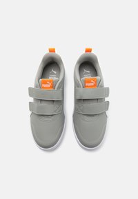 Baskets grises avec deux bandes Velcro, étiquette logo orange et semelle blanche. Matériau synthétique lisse avec des accents cousus sur la zone des orteils.
