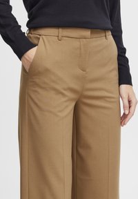 Hellbraune maßgeschneiderte Hose mit Seitentaschen, geraden Beinen und einer glatten Textur. Getragen mit einem schwarzen Langarmoberteil, das ein minimales Design zeigt.