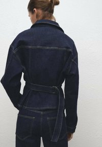 Chaqueta de denim con cintura ceñida, color azul oscuro, gran cuello, detalles de costura visibles y diseño de bolsillo trasero.