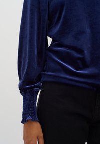 Tyrkysová sametová blůza s dlouhými rukávy, která má na manžetách nařasené detaily a hladkou strukturu. V kombinaci s černými kalhotami.