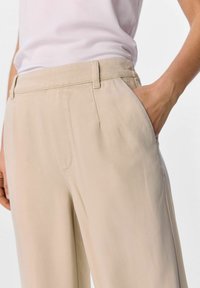 Lys beige bukser med en glat tekstur, der har sidelommer, en skræddersyet pasform og syet detaljer langs sømmene.