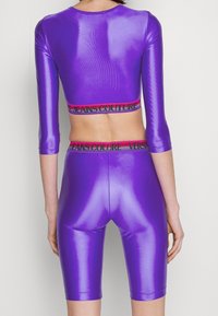 Purple glossy two-piece outfit consisting of a cropped top with three-quarter sleeves and matching high-waisted shorts, featuring logo bands.