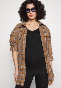 Chemise oversize à carreaux marron avec des manches roulées, poches avant, portée sur un haut noir, complétée par de grandes lunettes de soleil noires.