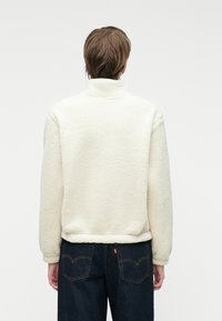 Pullover in pile color crema con colletto alto, superficie texture e orlo elastico, indossato con jeans blu scuro caratterizzati da cuciture sui taschini posteriori.