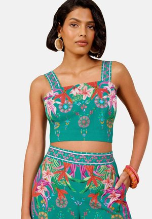 Vrouw draagt groen bloemig crop top en bijpassende broek, met grote ronde oorbellen en oranje armbanden om de pols.