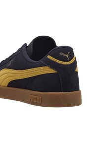 Baskets Puma discrètes en daim noir avec une bande moutarde, semelle en caoutchouc et logo Puma au talon et sur le côté de la semelle.