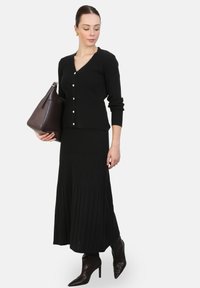 Cardigan noir en tricot côtelé avec décolleté en V et détails de boutons, associé à une jupe midi plissée noire. Les accessoires comprennent un sac à main marron foncé et des bottes à talons.