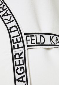 Tissu blanc à finition texturée, avec une bande élastique noire proéminente affichant "KARL LAGERFELD." Design distinctif avec lettres contrastantes.
