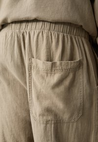 Khaki színű pamut nadrág, elasztikus derékpánttal, egy hátsó zsebbel és puha, texturált anyaggal. Finom gyűrődések láthatók.