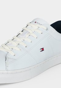 Sneaker in pelle bianca con lacci bianchi, con un piccolo logo Tommy Hilfiger rosso, bianco e blu sul lato e linguetta con marchio.